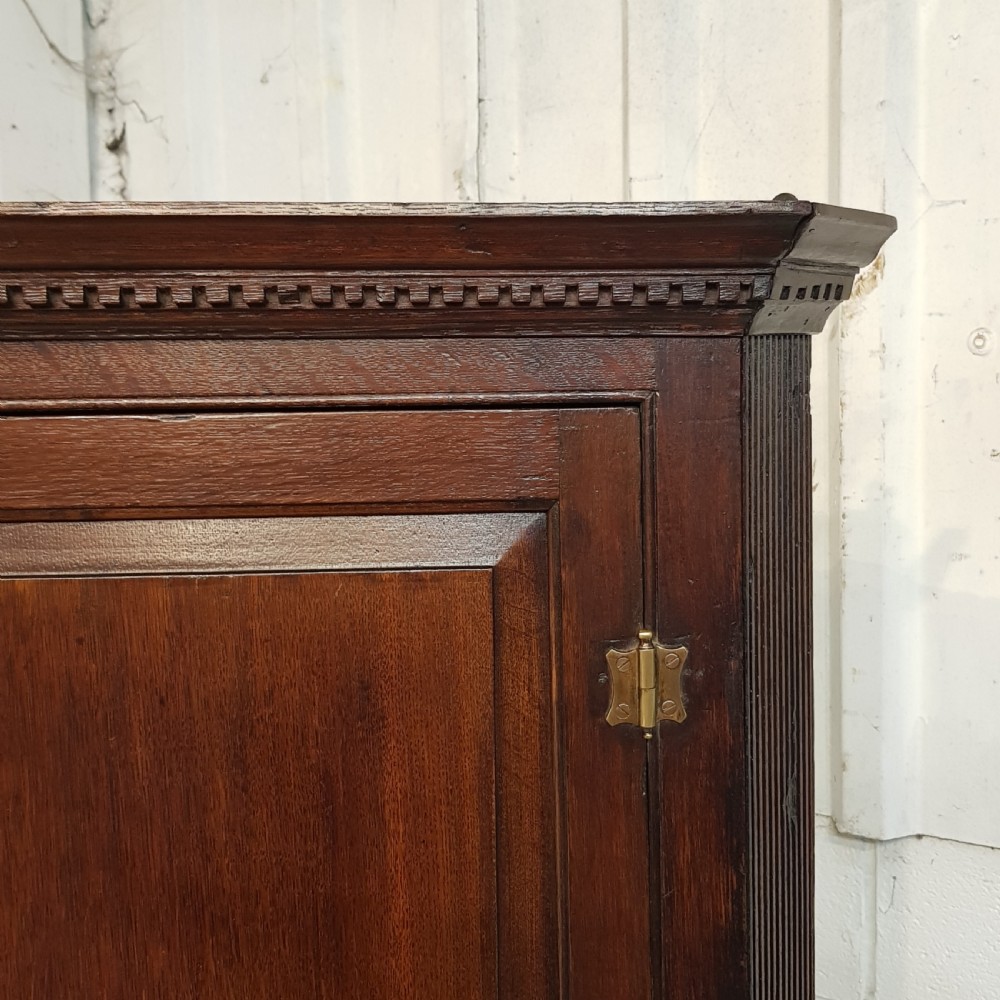 An 18th-century dark oak wall-hanging corner cabinet featuring a single panelled door with brass H-hinges and a keyhole escutcheon, topped with a dentil moulded cornice. View VI