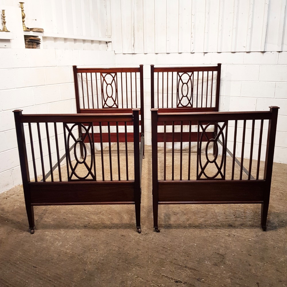 A pair of Edwardian mahogany single bed frames featuring slatted headboards and footboards accented with central geometric oval motifs and standing on castors.