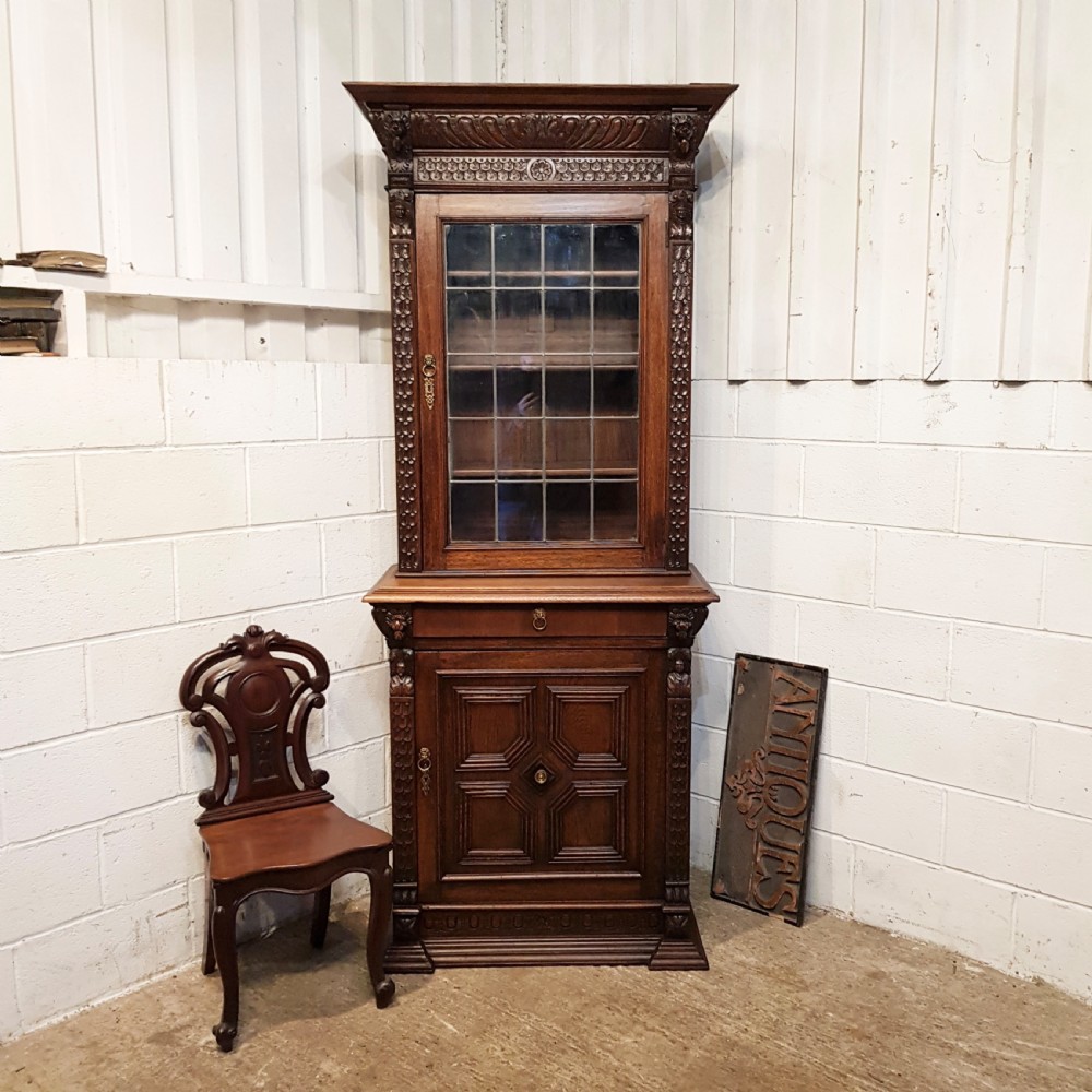 A tall, narrow Victorian Gothic carved oak bookcase cabinet featuring a single glazed door above a geometric panelled lower cupboard door.