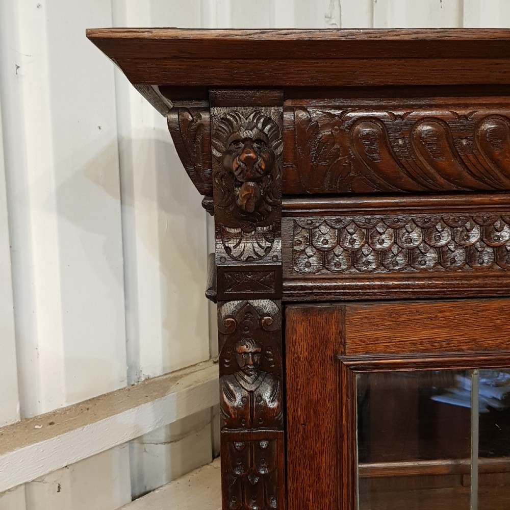A tall, narrow Victorian Gothic carved oak bookcase cabinet featuring a single glazed door above a geometric panelled lower cupboard door. View II