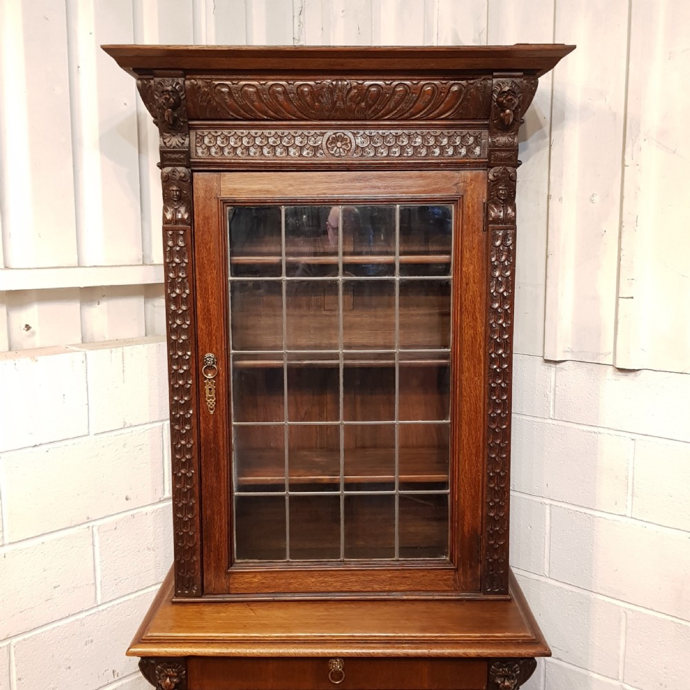 A tall, narrow Victorian Gothic carved oak bookcase cabinet featuring a single glazed door above a geometric panelled lower cupboard door. View III