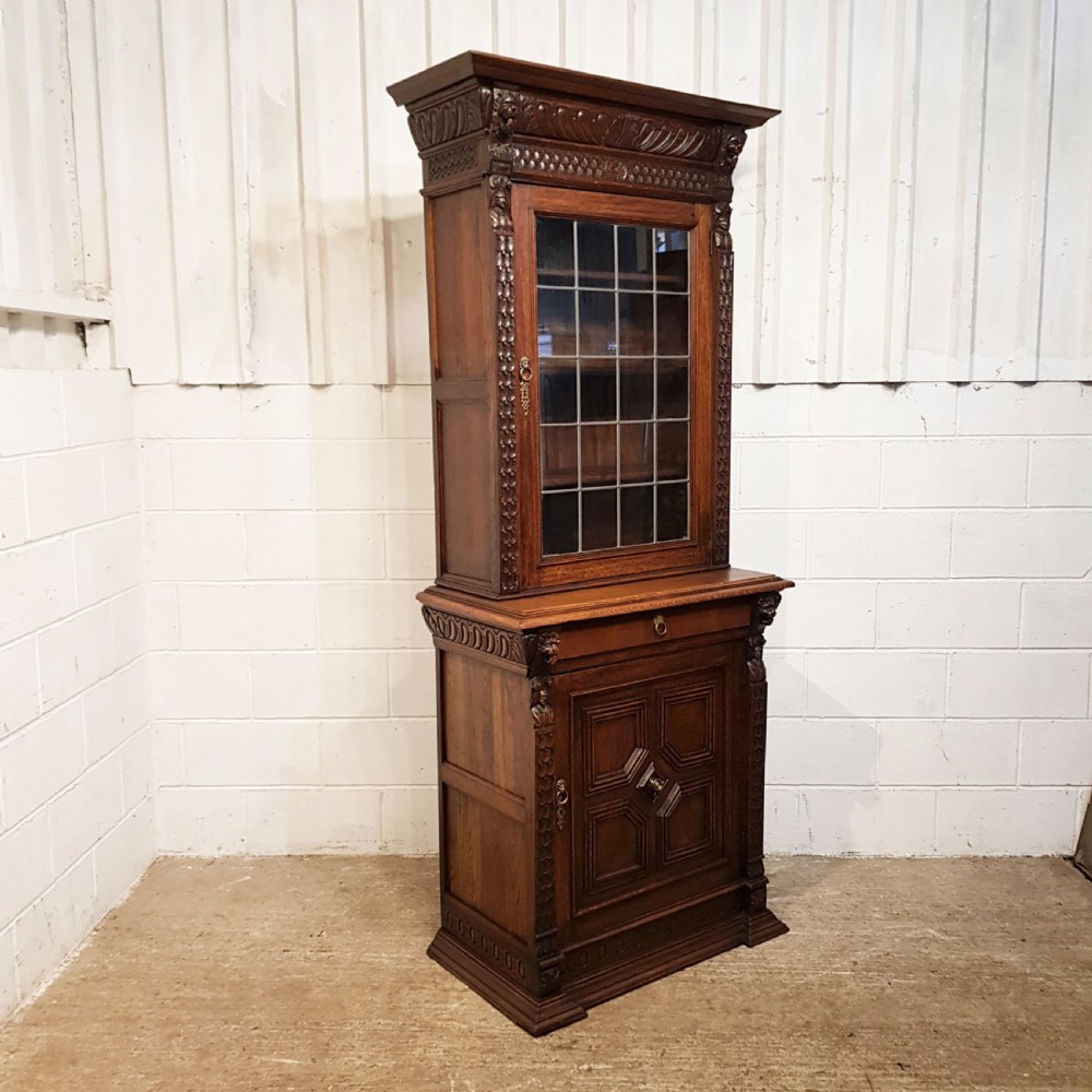 A tall, narrow Victorian Gothic carved oak bookcase cabinet featuring a single glazed door above a geometric panelled lower cupboard door. View VI