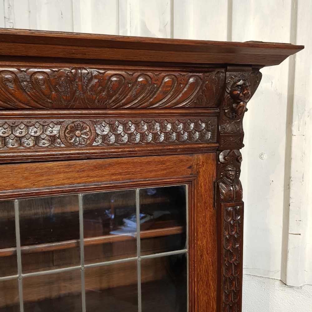 A tall, narrow Victorian Gothic carved oak bookcase cabinet featuring a single glazed door above a geometric panelled lower cupboard door. View VIII