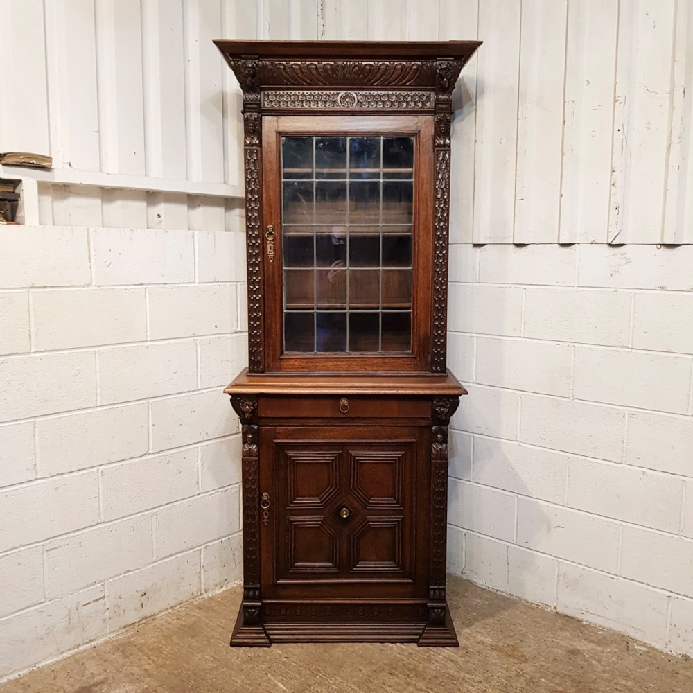 A tall, narrow Victorian Gothic carved oak bookcase cabinet featuring a single glazed door above a geometric panelled lower cupboard door. View X