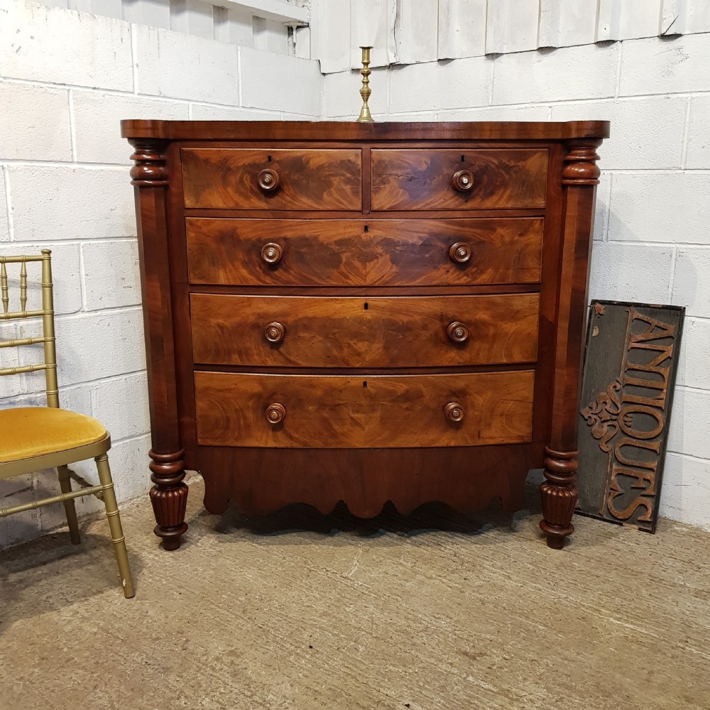 An antique Victorian bow-fronted Scotch chest of drawers featuring flame mahogany veneer, turned side columns, and turned wooden knobs.