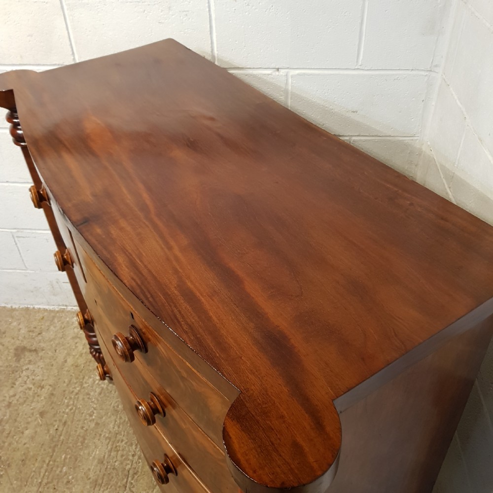 An antique Victorian bow-fronted Scotch chest of drawers featuring flame mahogany veneer, turned side columns, and turned wooden knobs. View V