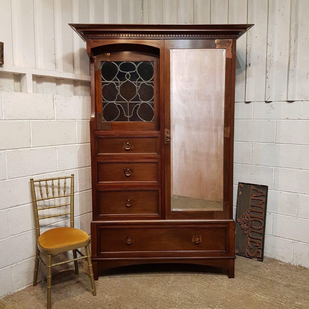 An antique Arts and Crafts mahogany combination wardrobe featuring a leaded glass cabinet, multiple drawers, copper strap hinges, and a tall mirrored door.