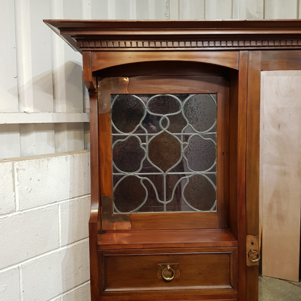 An antique Arts and Crafts mahogany combination wardrobe featuring a leaded glass cabinet, multiple drawers, copper strap hinges, and a tall mirrored door. View II