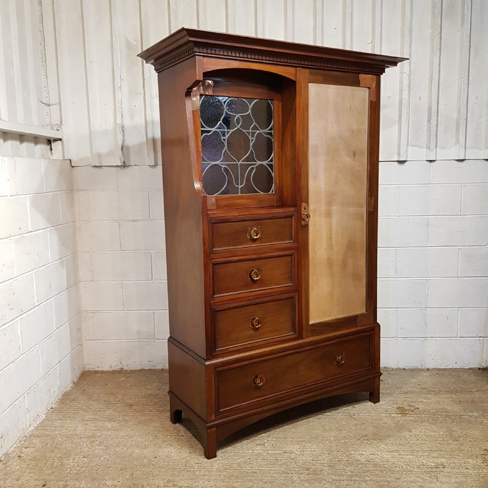 An antique Arts and Crafts mahogany combination wardrobe featuring a leaded glass cabinet, multiple drawers, copper strap hinges, and a tall mirrored door. View VI