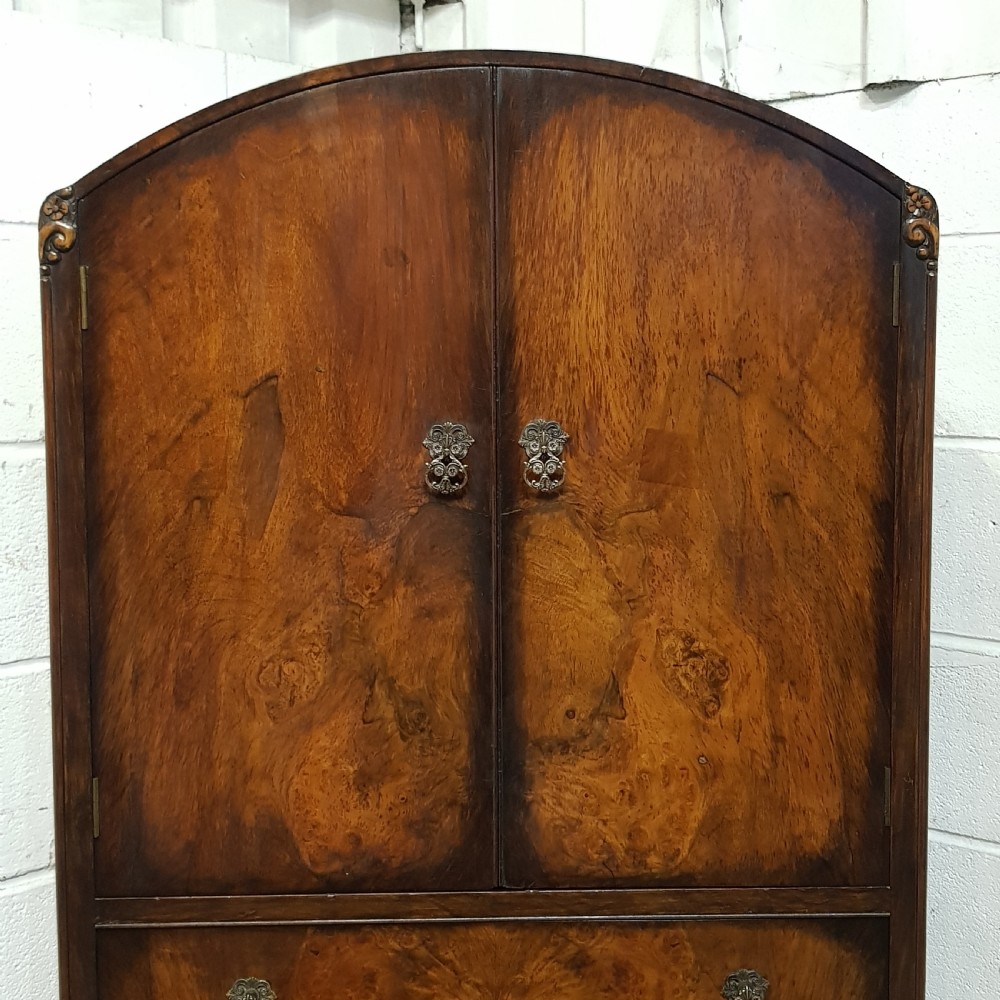 A 1920s burr walnut tallboy featuring an arched top two-door cabinet over three wide drawers with ornamental brass handles and cabriole feet. View VI