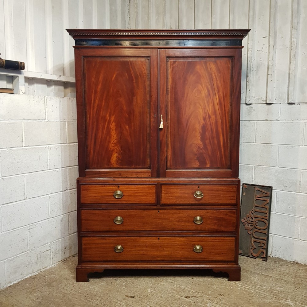 A George III mahogany clothes press featuring flame mahogany panelled upper doors above a base with two short and two long drawers on bracket feet.