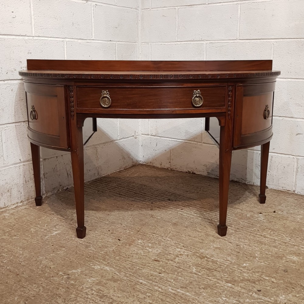An antique mahogany demi-lune side desk featuring a curved front, central drawer with brass ring handles, and tapered square legs.