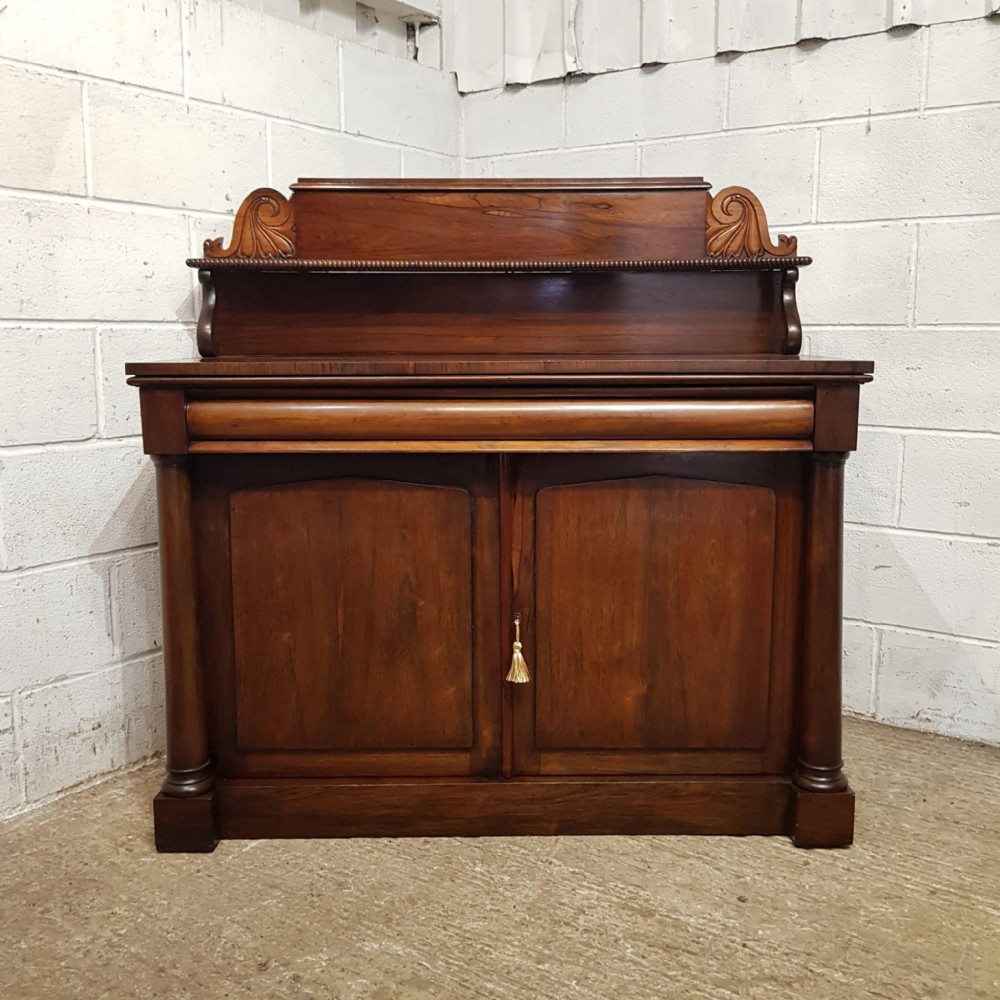 An Early Victorian dark rosewood chiffonier featuring a raised shelf upstand with scroll carvings, a cushion drawer, and a two-door lower cabinet flanked by turned columns.