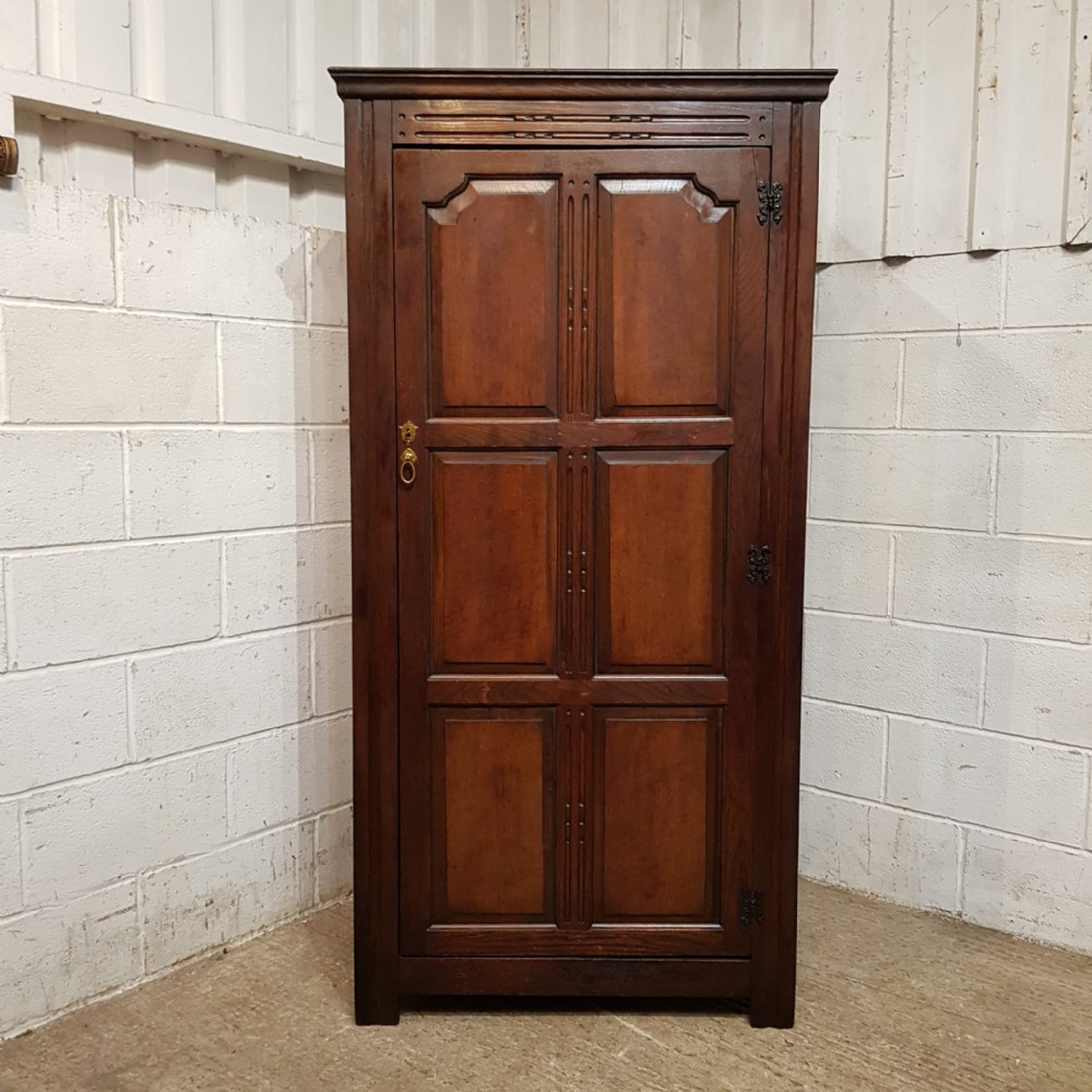 A compact dark oak single-door wardrobe dating to circa 1920, featuring six recessed panels, carved detailing, and exposed decorative hinges.