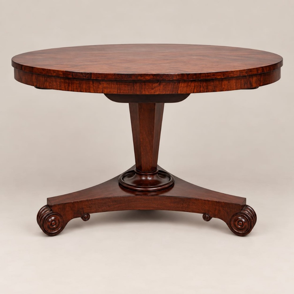An antique circular William IV rosewood tilt-top dining table with a polished surface, central column stem, and triform plinth base with carved scroll feet.