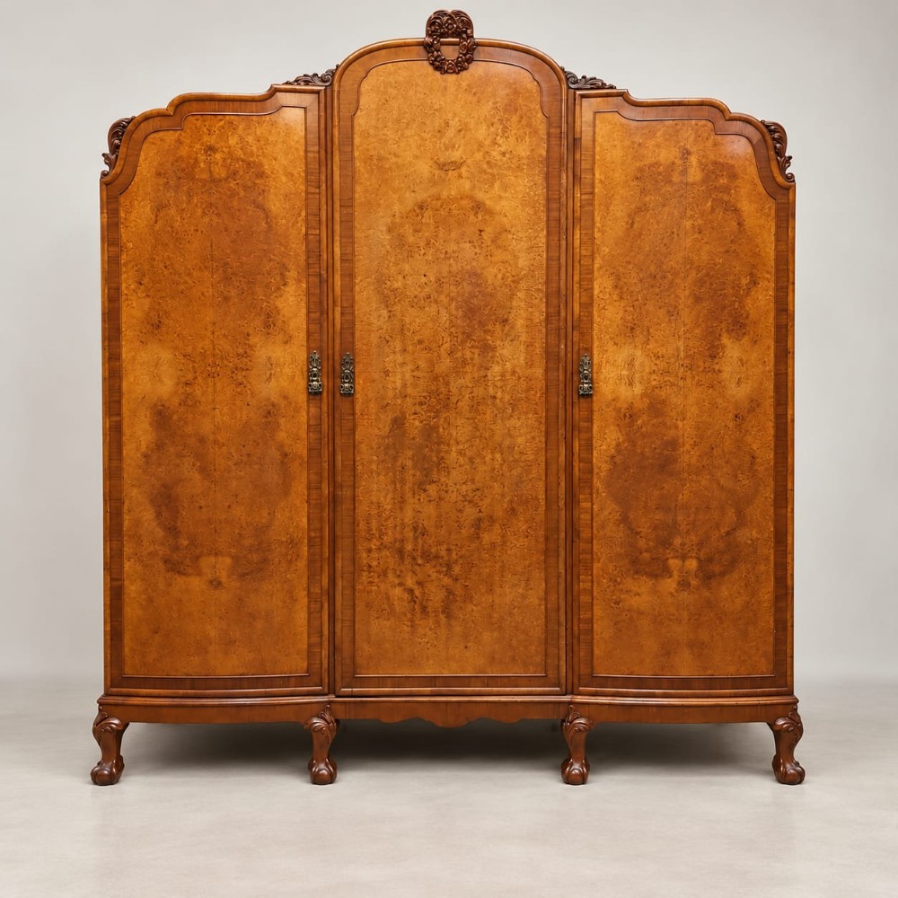 An antique circa 1920 burr walnut triple wardrobe featuring three panelled doors with figured wood grain, carved top cresting, and carved cabriole feet.