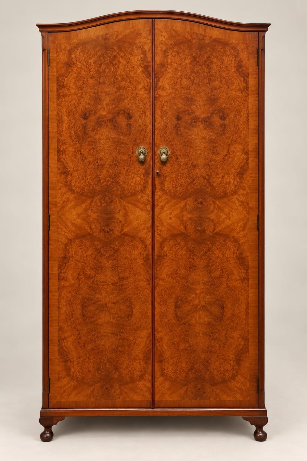 A two-door 1920s small burr walnut wardrobe featuring symmetrical bookmatched wood grain, a gently arched top, brass drop handles, and short turned feet.