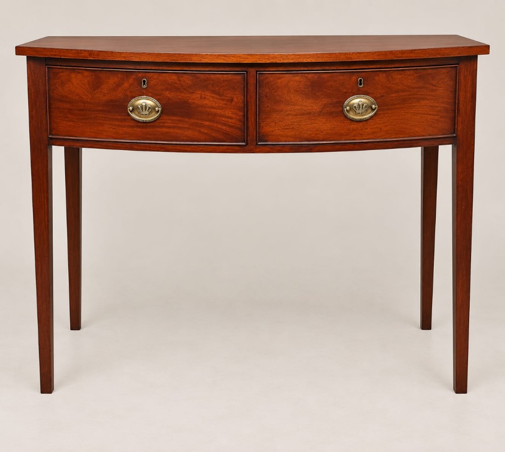 A Regency mahogany bow-fronted side table featuring two drawers with brass handles and supported by four tapered legs.