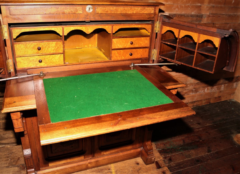 A Victorian walnut and maple cylinder desk featuring curved upper shutters, burr walnut panel inlays, a carved pediment, and double cabinet doors below. View III