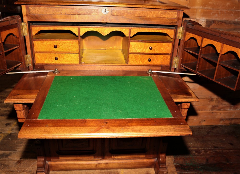 A Victorian walnut and maple cylinder desk featuring curved upper shutters, burr walnut panel inlays, a carved pediment, and double cabinet doors below. View V