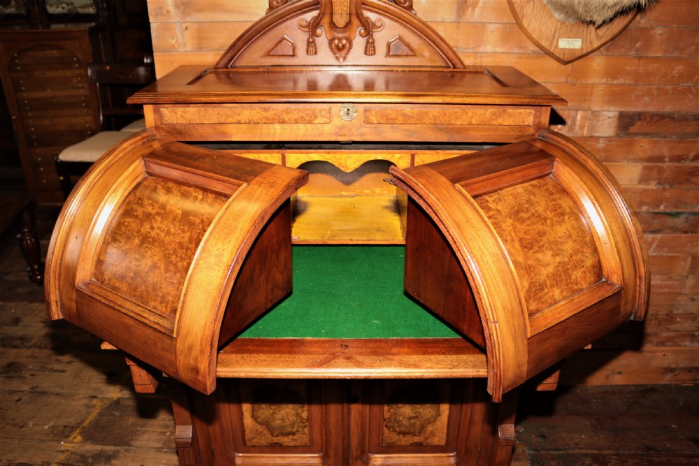 A Victorian walnut and maple cylinder desk featuring curved upper shutters, burr walnut panel inlays, a carved pediment, and double cabinet doors below. View VIII