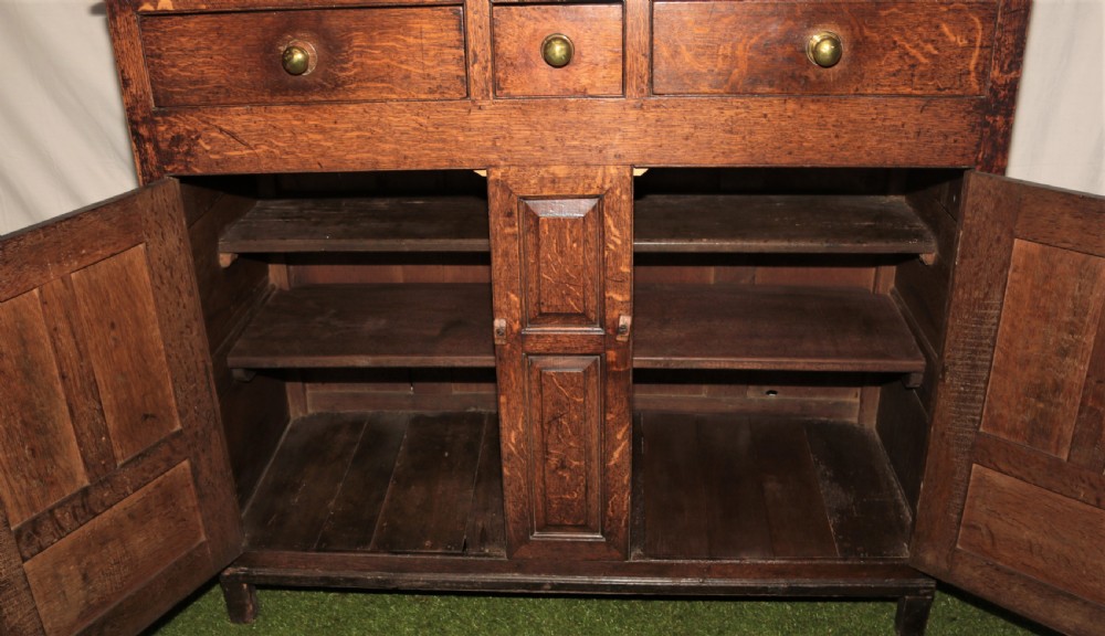 A dark oak court cupboard featuring arched panelled doors, drop finials, three middle drawers, and brass knobs. View III