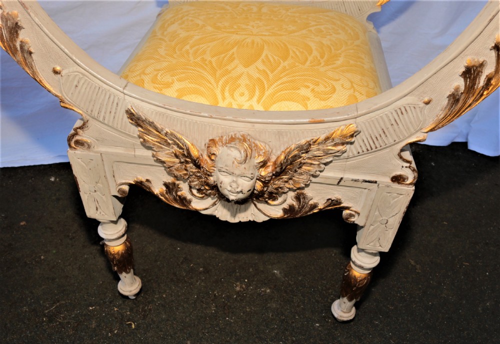 A pair of painted and carved giltwood Savonarola chairs with curved X-frames, ornate carved crests, and yellow patterned fabric seat cushions. View VIII