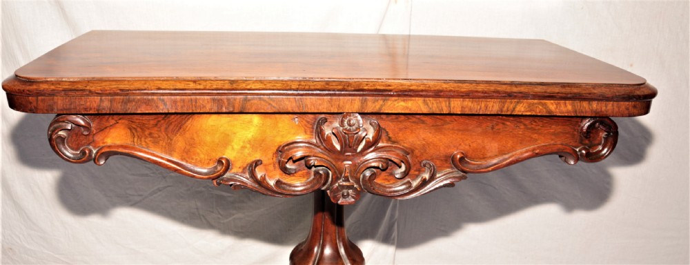 A William IV rosewood fold-over games table featuring an ornate carved apron, a sculpted central pedestal, and a four-legged carved base with brass castors. View V