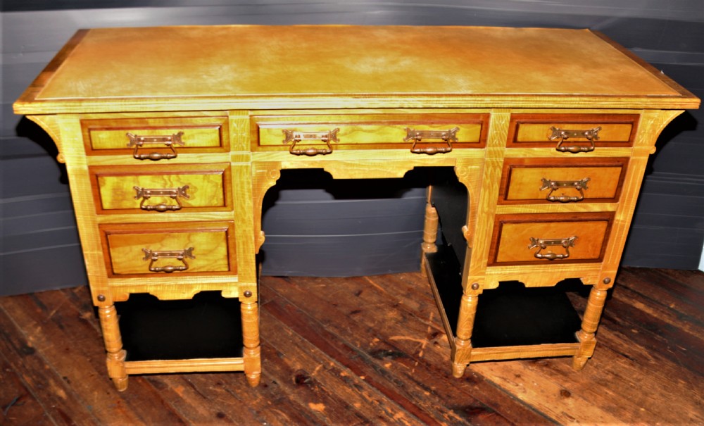 An Arts and Crafts light ash pedestal desk featuring a leather writing surface, seven drawers with brass handles, and open lower shelves.