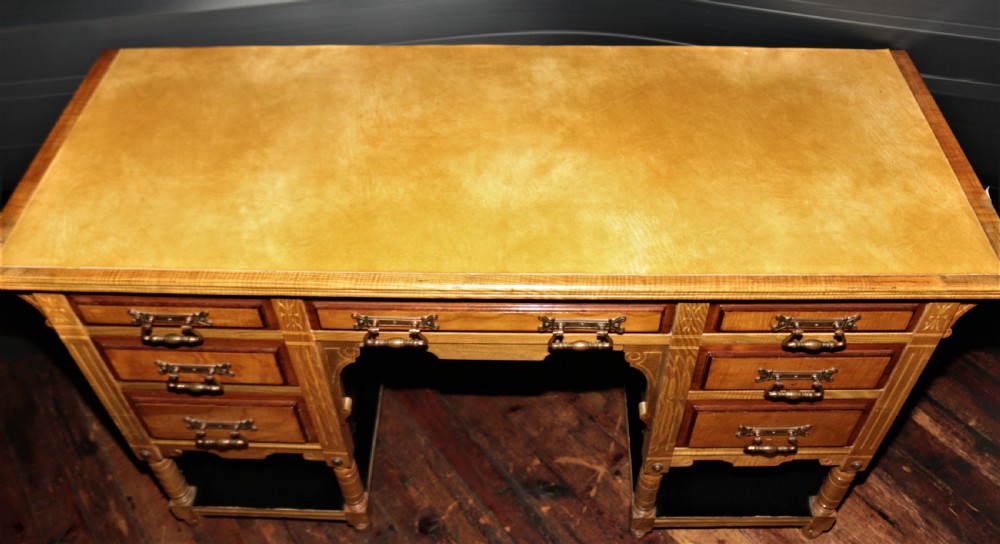 An Arts and Crafts light ash pedestal desk featuring a leather writing surface, seven drawers with brass handles, and open lower shelves. View V