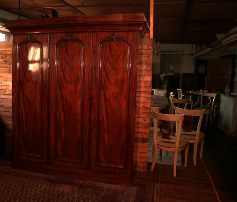 A large Victorian mahogany triple wardrobe featuring three arched panel doors with flame mahogany veneer figure, carved scroll crests, and a heavy moulded cornice. View V