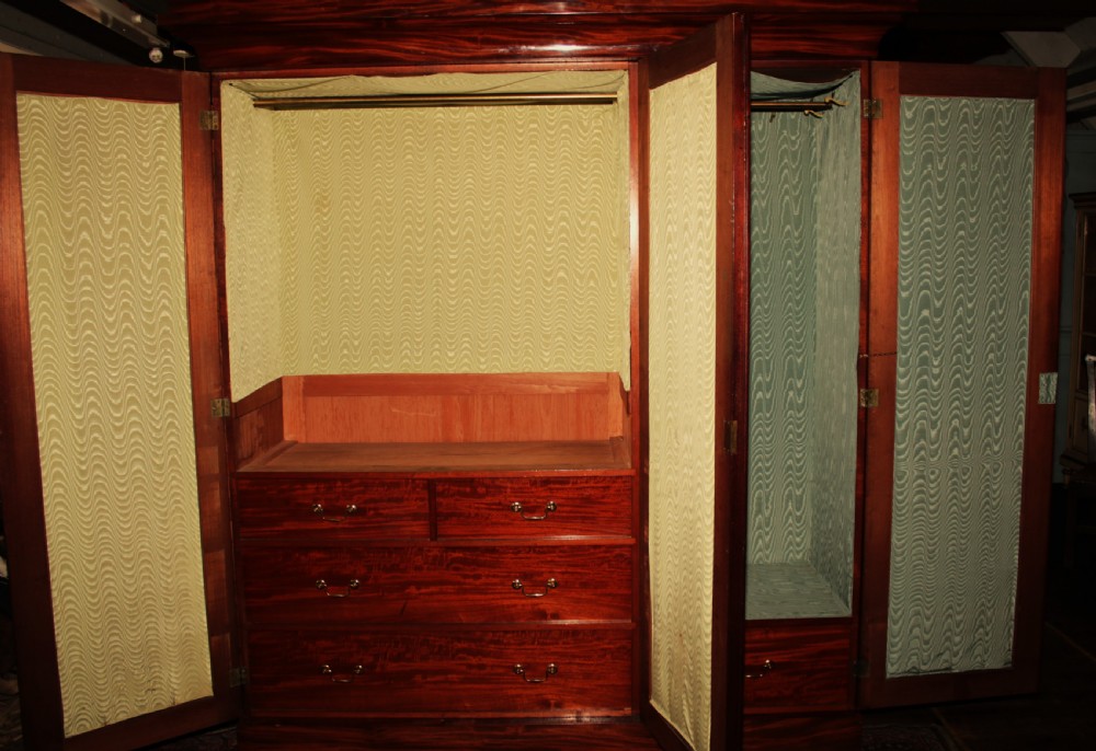 A large Victorian mahogany triple wardrobe featuring three arched panel doors with flame mahogany veneer figure, carved scroll crests, and a heavy moulded cornice. View VII