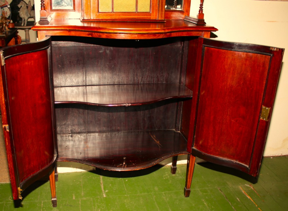 An Edwardian inlaid mahogany display cabinet featuring a central glazed door with Gothic tracery, mirrored side panels, a broken pediment cresting, and a serpentine lower cupboard. View V