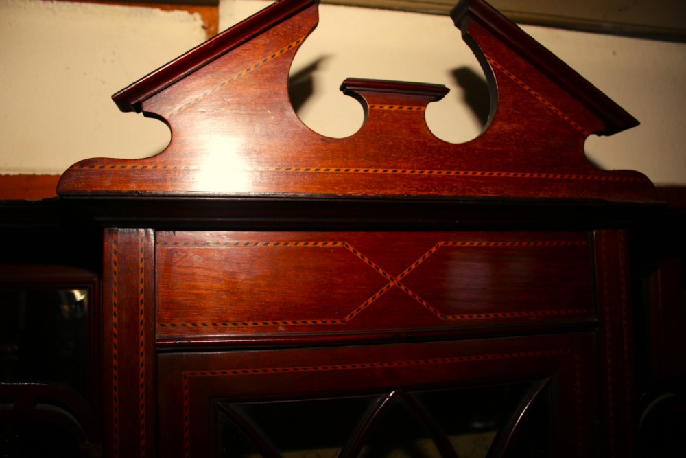 An Edwardian inlaid mahogany display cabinet featuring a central glazed door with Gothic tracery, mirrored side panels, a broken pediment cresting, and a serpentine lower cupboard. View VII