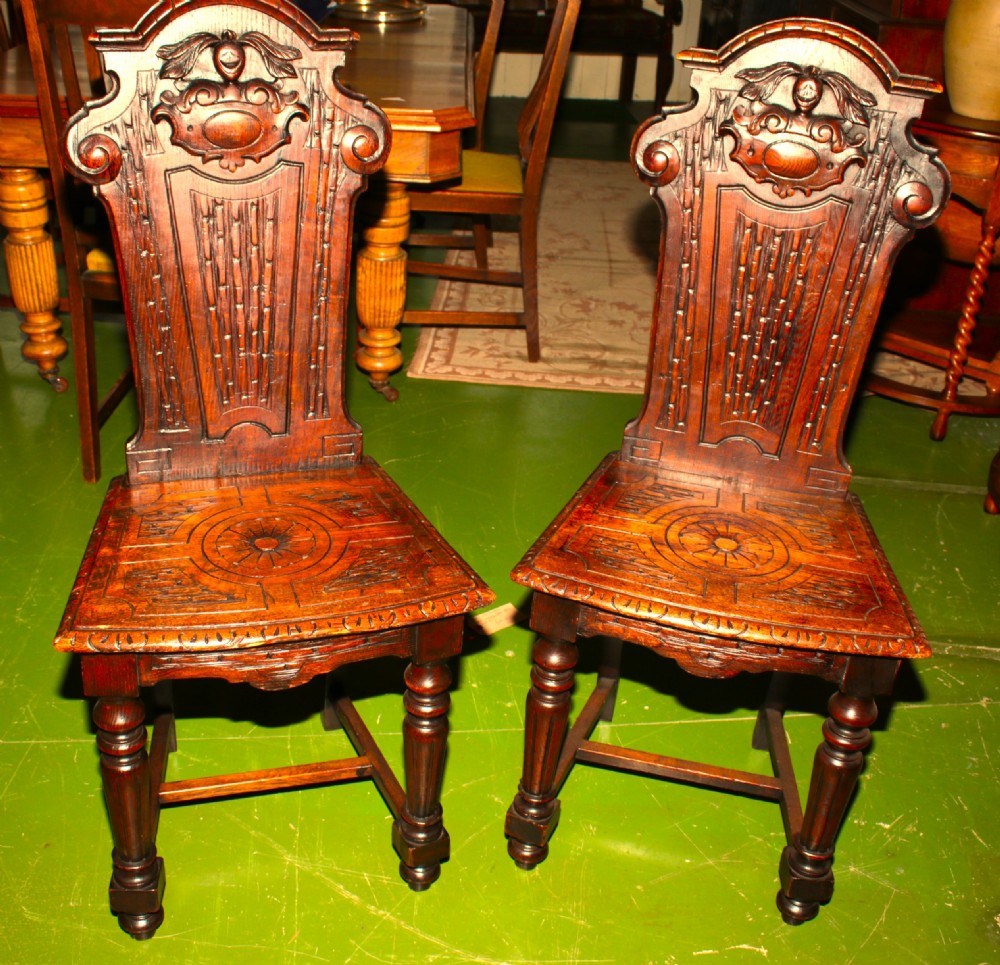 A pair of carved dark oak hall chairs featuring ornate back panels, carved wooden seats, and turned front legs.