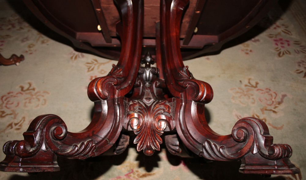 A Victorian rosewood tilt-top dining table set vertically to reveal its dark figured grain pattern and scalloped serpentine top resting on a carved tripod base. View II