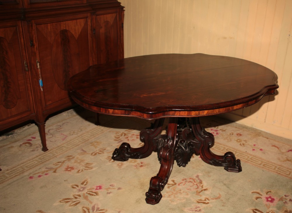 A Victorian rosewood tilt-top dining table set vertically to reveal its dark figured grain pattern and scalloped serpentine top resting on a carved tripod base. View IX