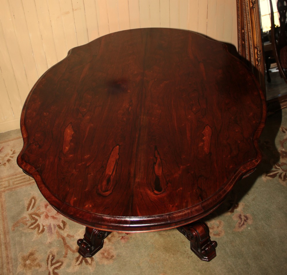 A Victorian rosewood tilt-top dining table set vertically to reveal its dark figured grain pattern and scalloped serpentine top resting on a carved tripod base. View X