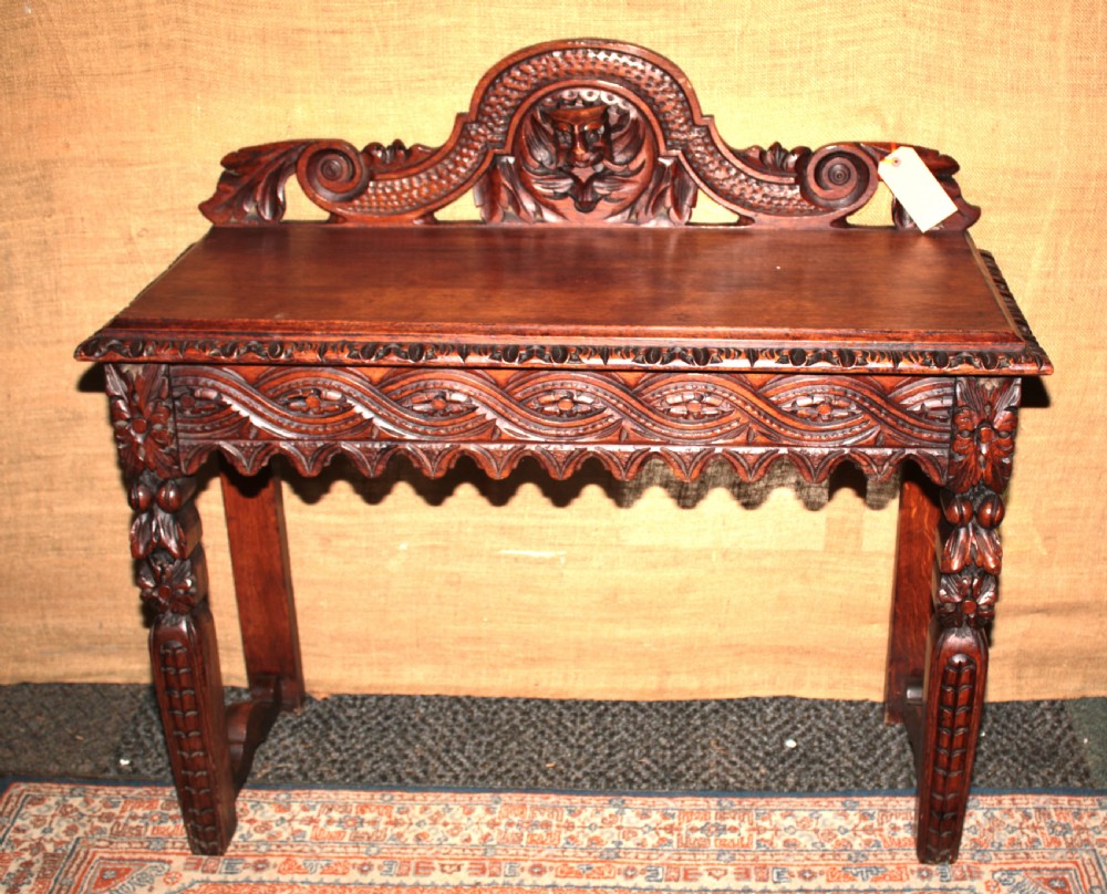 A dark carved oak side table featuring an ornate upstand with a mask motif, an intricately carved apron, and detailed legs.
