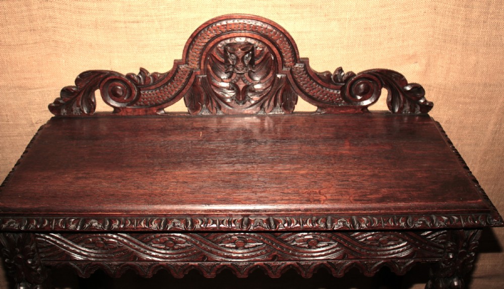 A dark carved oak side table featuring an ornate upstand with a mask motif, an intricately carved apron, and detailed legs. View VII
