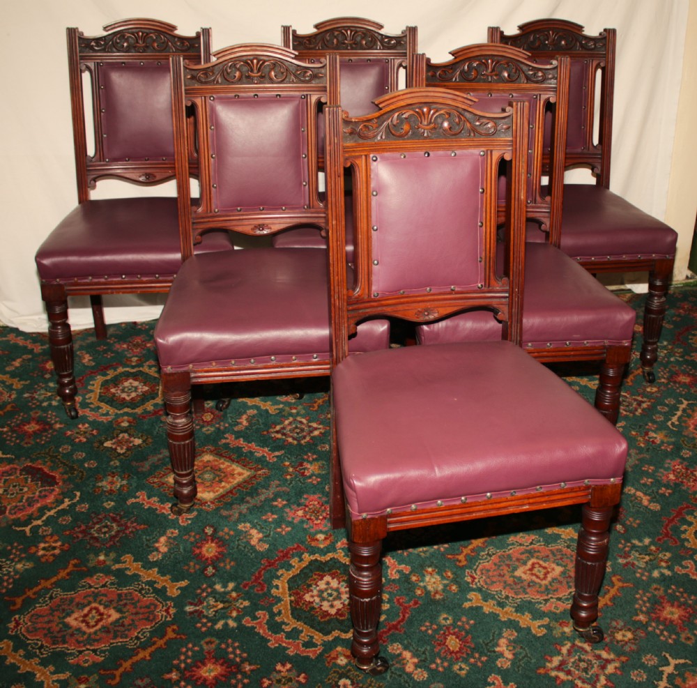 A set of six Victorian Irish mahogany dining chairs upholstered in deep plum leather with brass stud detailing, carved crests, and fluted front legs on castors.
