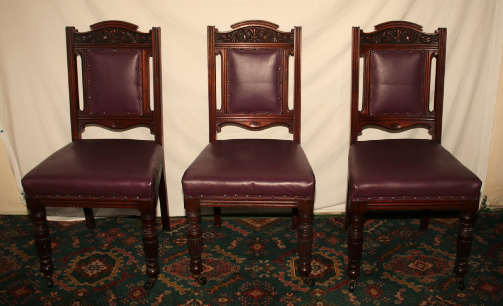 A set of six Victorian Irish mahogany dining chairs upholstered in deep plum leather with brass stud detailing, carved crests, and fluted front legs on castors. View II