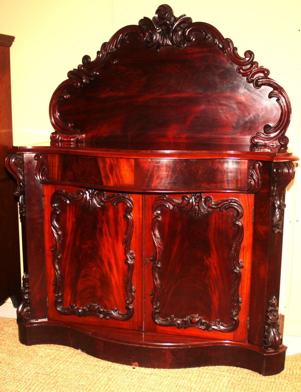 A Victorian mahogany two-door sideboard featuring a carved scrollwork backboard, flame mahogany panels, and a serpentine plinth base.
