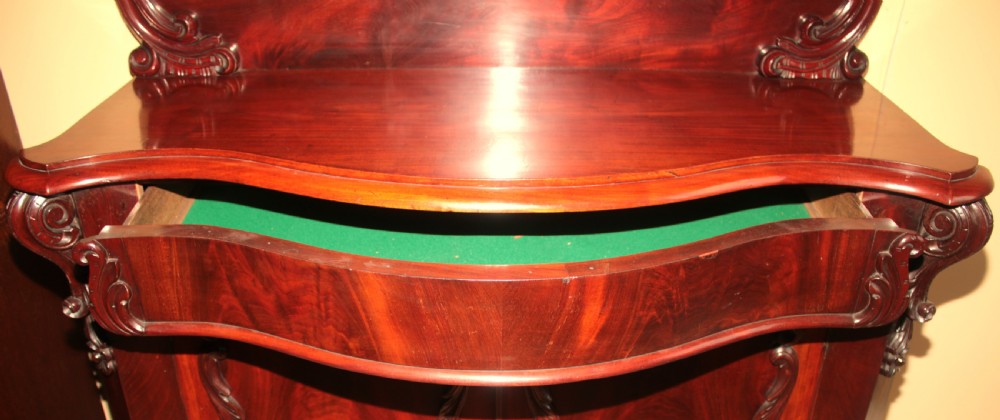 A Victorian mahogany two-door sideboard featuring a carved scrollwork backboard, flame mahogany panels, and a serpentine plinth base. View VI