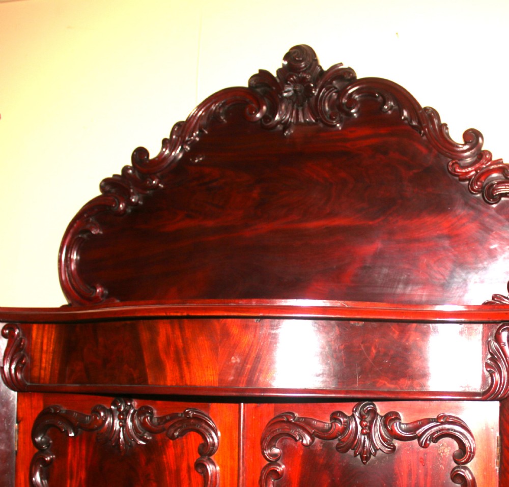 A Victorian mahogany two-door sideboard featuring a carved scrollwork backboard, flame mahogany panels, and a serpentine plinth base. View X