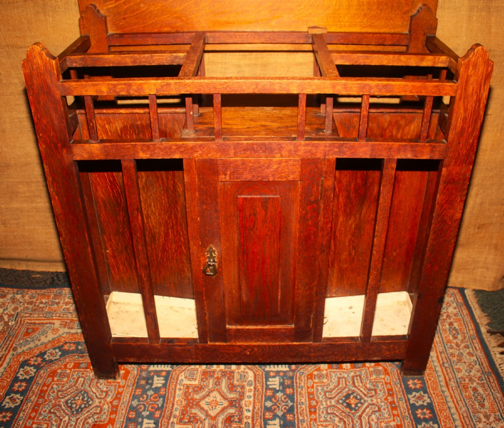 An Arts and Crafts oak hall stand featuring a central square mirror, metal coat hooks, umbrella drip trays, and a lower storage cupboard. View II
