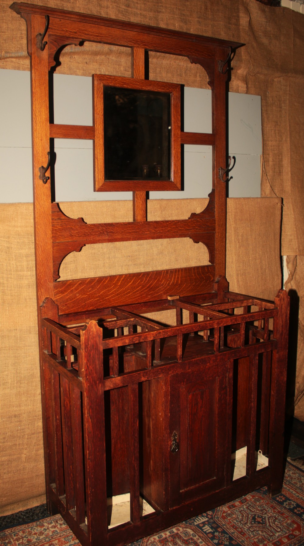 An Arts and Crafts oak hall stand featuring a central square mirror, metal coat hooks, umbrella drip trays, and a lower storage cupboard. View IV