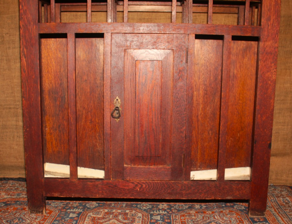 An Arts and Crafts oak hall stand featuring a central square mirror, metal coat hooks, umbrella drip trays, and a lower storage cupboard. View VII