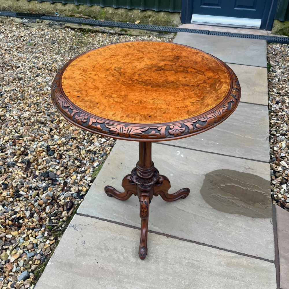A round Victorian occasional table featuring a figured burr walnut top with carved edge detailing, supported by a central turned pedestal and three scrolled legs.