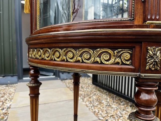 An oval French mahogany vitrine featuring glass side panels, gilt-bronze ormolu mounts, fluted legs, and a gallery top above a lower undertier shelf. View V